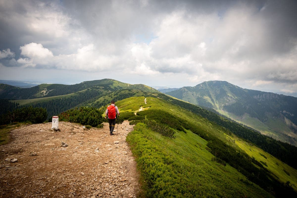 A hiker on a trail through green mountains under a cloudy sky