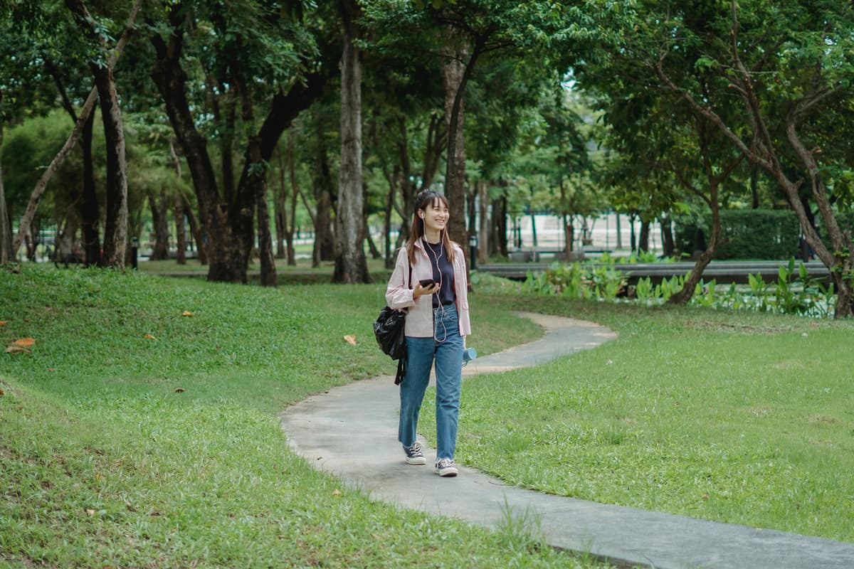 A woman walking along a park path with earphones in, holding her phone